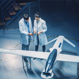 Two people in white lab coats speaking in a lab near a model plane.