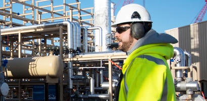 A person wearing a reflective jacket and safety equipment stands next to a system of pipes.

