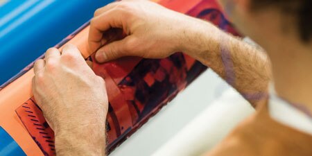 Close-up of hands carefully applying a red printing plate to a mounting cylinder.
