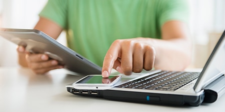 a person holding a tablet while tapping the screen of a smart phone which rests on a laptop.
