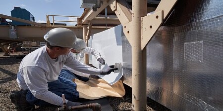 Two people wearing hard hats and gloves apply a roll of insulating material to siding.
