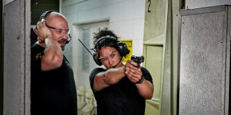 Two individuals at an indoor shooting range, with one person instructing the other on how to properly hold and aim a handgun. Both are wearing protective ear gear.
