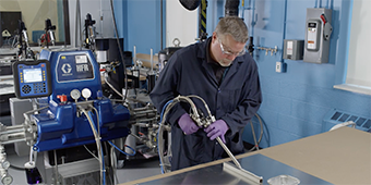 Video still of a worker applying adhesive at a bench using a powered applicator
