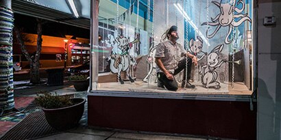 Man kneeling in retail display window with drawn characters in the display.
