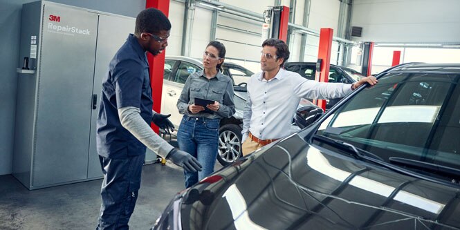 Three people talk and gesture while standing next to a car in an autobody shop.

