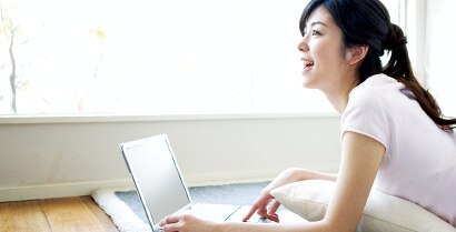 A young woman happily works on a laptop near a sunny window, unconcerned about glare.
